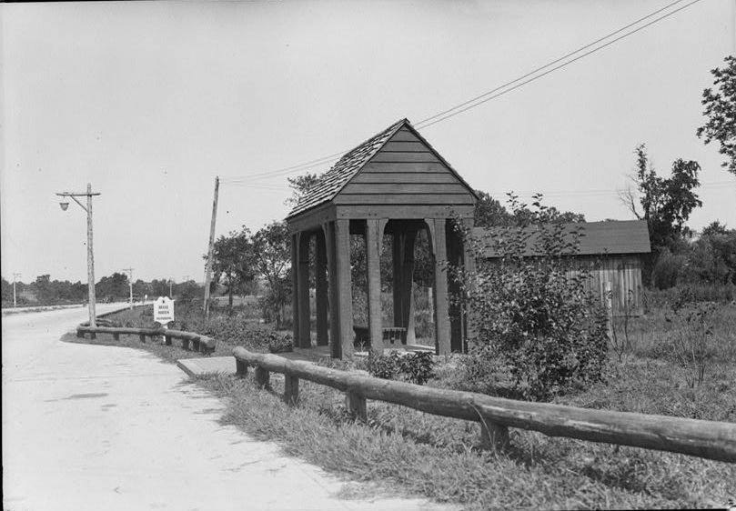 A small open-sided wooden shelter with a shingled roof beside the new parkway, with a Belle Haven sign and log guardrails.