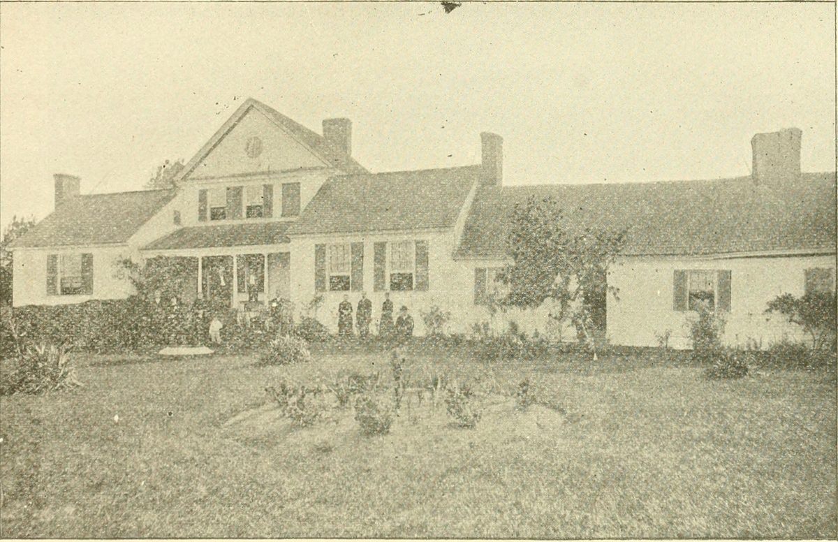A long white farmhouse with chimneys at each end, people standing in front, about 1904.