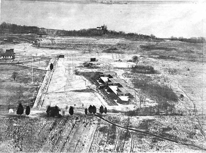 Aerial view over cleared ground toward the Potomac, with low battery buildings in the center and a line of trees in the foreground.