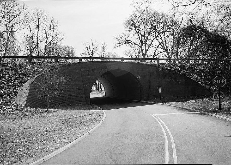 A brick-faced concrete arch carrying the parkway over a curving road, with bare trees and a wooden guardrail above.