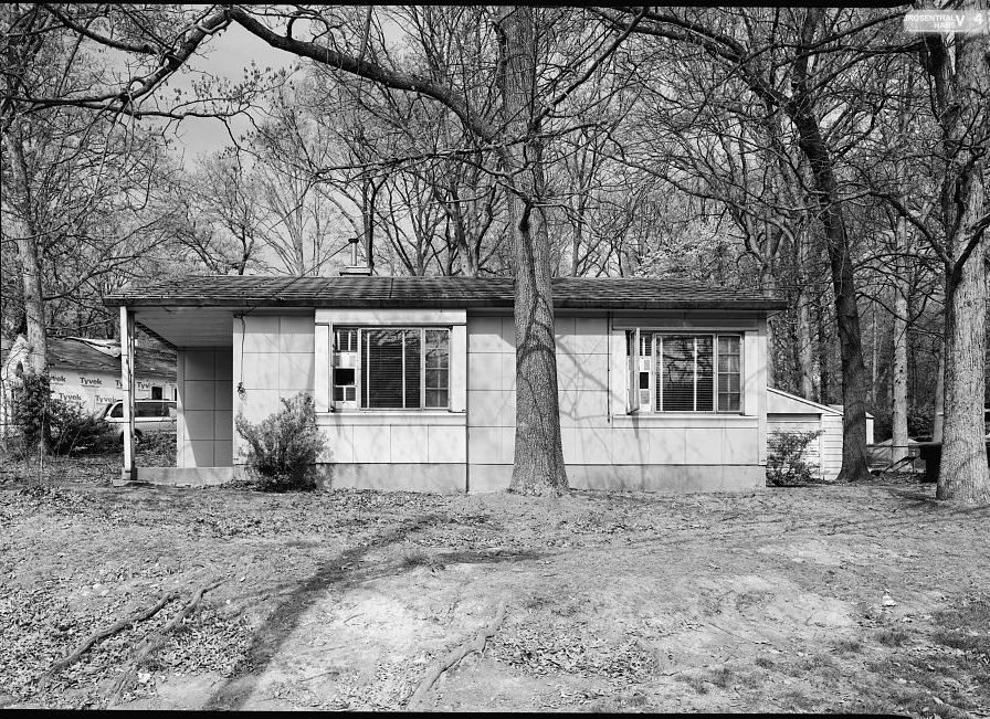 A one-story Lustron house with walls of square enameled metal panels, under tall trees.
