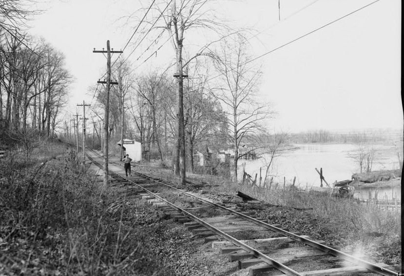 Single-track electric railway beside the Potomac, with poles and wires, a small station shed, and one man walking up the line.